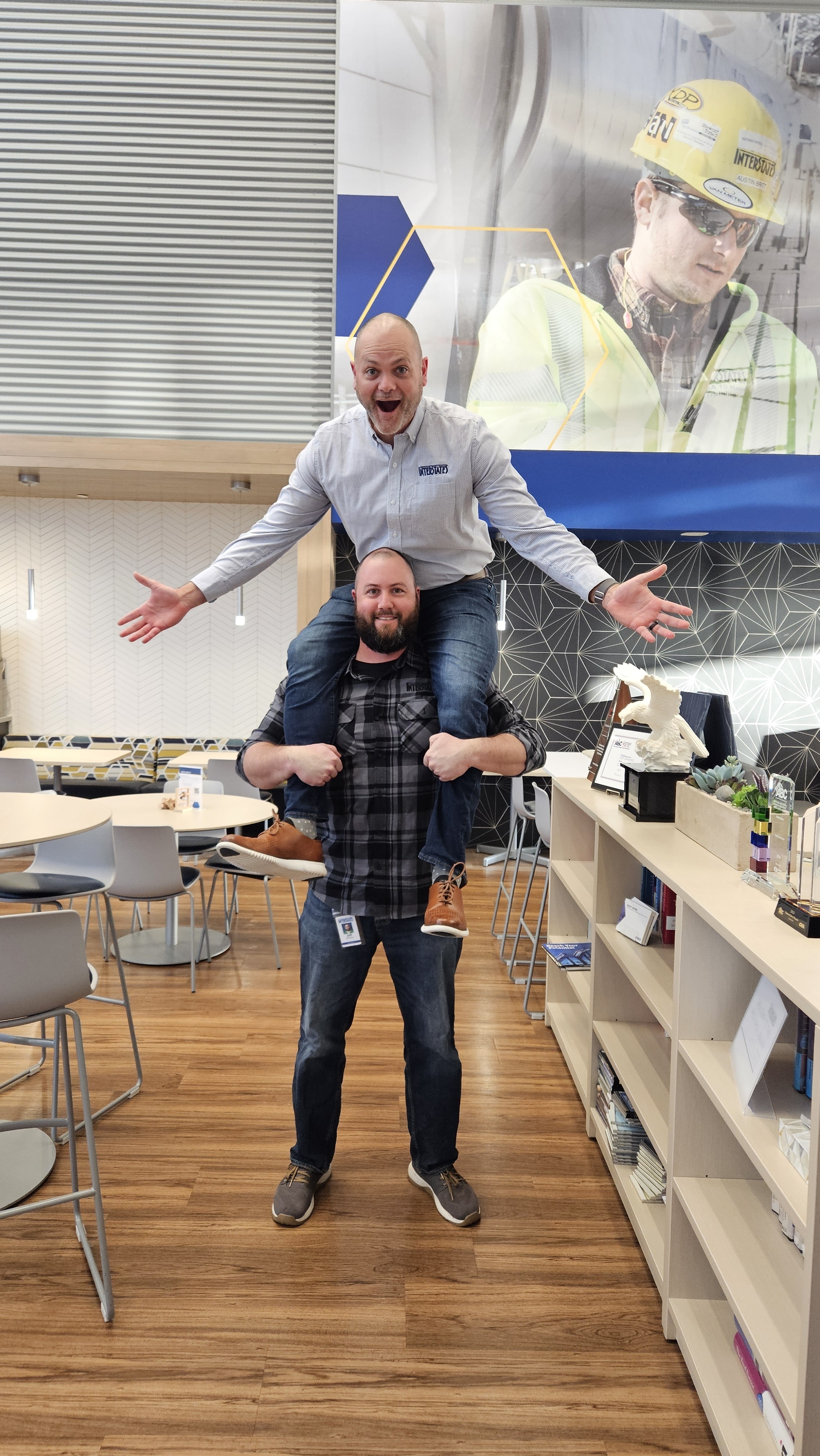 Playful office moment with one employee sitting on another's shoulders in break room, surrounded by modern décor and company branding
