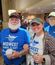 A smiling veteran and a young woman stand arm in arm indoors, both wearing “Midwest Honor Flight” shirts — his blue and hers white — along with hats. The veteran holds a cup, and the woman holds a can, surrounded by a group of others in similar Honor Flight shirts in the background. The setting appears to be a hotel lobby with warm lighting and people preparing for an event.