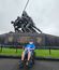 A veteran in a blue “Midwest Honor Flight” shirt sits smiling in a wheelchair in front of the United States Marine Corps War Memorial in Arlington, Virginia. The bronze statue behind him depicts six Marines raising the American flag on Iwo Jima, with the flag waving above them against a cloudy gray sky. The memorial’s base features gold lettering honoring fallen Marines.