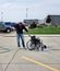 A person wearing jeans and a navy Interstates sweatshirt uses a power washer to clean a wheelchair in a parking lot. Water sprays from the hose onto the pavement as the person leans slightly forward directing the stream. Several vehicles, a grassy area, and industrial buildings are visible in the background under a cloudy sky.