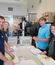 Four people stand around a countertop in an office breakroom, smiling as they fold and label envelopes. Postal bins labeled “United States Postal Service” are visible on the counter, along with stacks of papers and mail labels. The background includes kitchen appliances, cabinets, and a glass wall revealing another office area.
