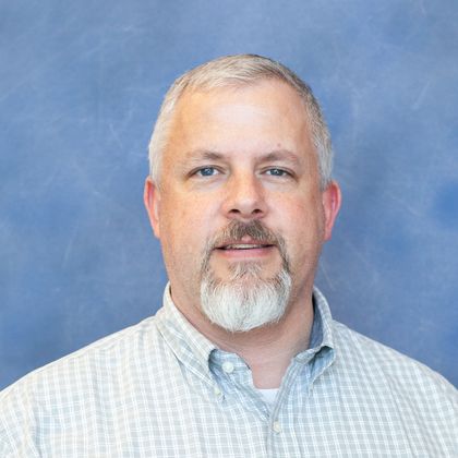 A head-and-shoulders portrait of a middle-aged man with short gray hair and a gray goatee, wearing a light blue checkered button-down shirt, posed against a blue studio backdrop.
