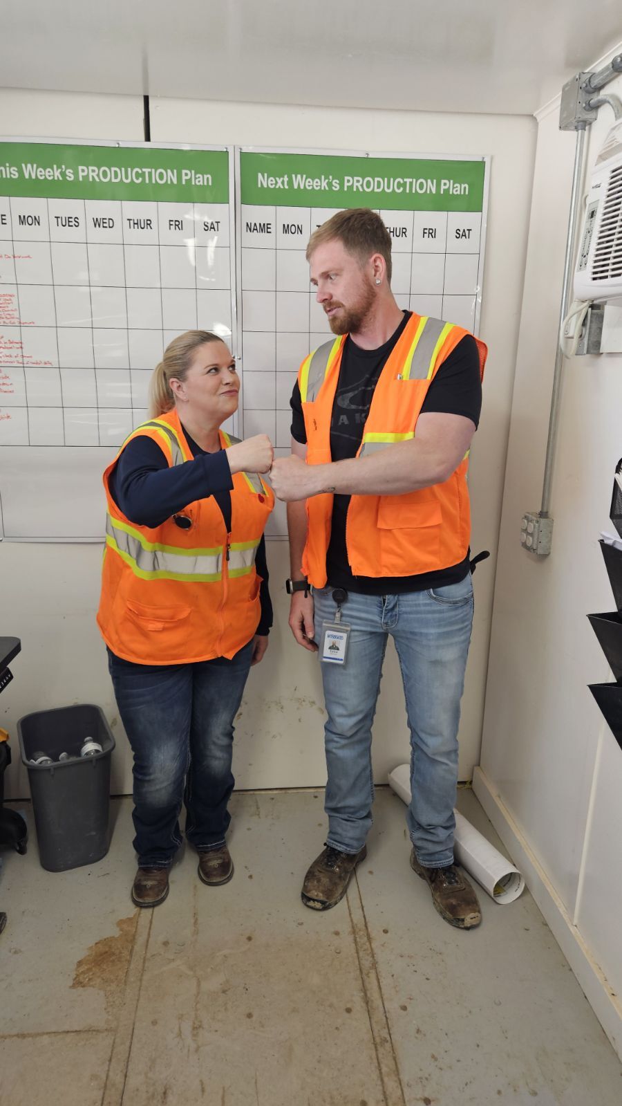 Two workers in safety vests discussing next week's production plan shown on wall-mounted schedule boards