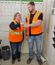 Two workers in safety vests discussing next week's production plan shown on wall-mounted schedule boards