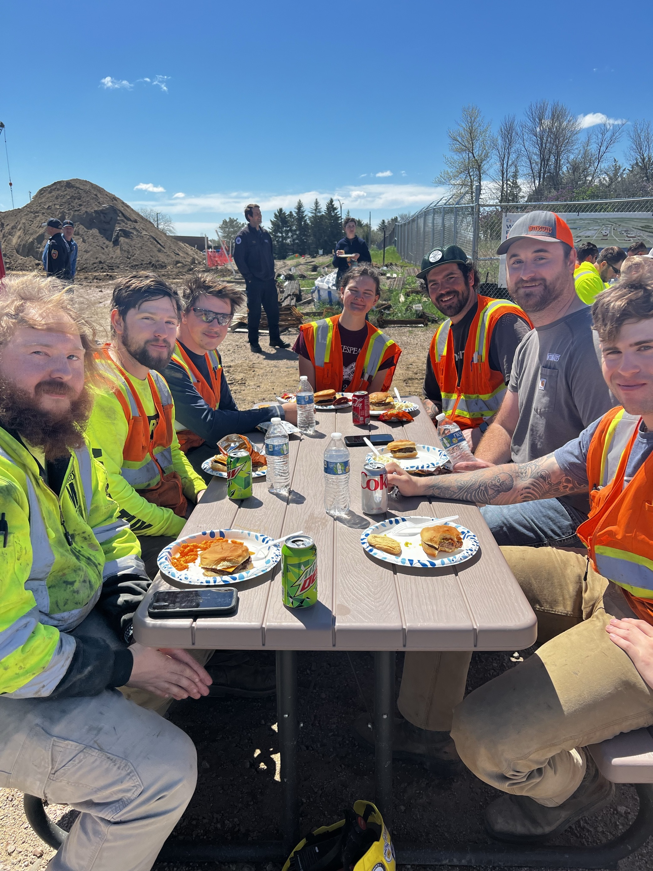Construction crew having lunch break outdoors at folding table, enjoying burgers and drinks while wearing safety vests, with dirt pile and fence visible in background