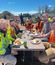 Construction crew having lunch break outdoors at folding table, enjoying burgers and drinks while wearing safety vests, with dirt pile and fence visible in background