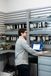 A technician stands at a workbench in an industrial controls lab, using a laptop in front of mounted electrical and automation components on wall racks.