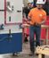 A man in an orange ABC shirt, white hard hat, gloves, and jeans walks past a training competition setup while holding a tape measure. Another person stands behind him near the booth.