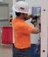 A man in an orange ABC shirt and white hard hat works on a piping assembly panel during a competition, using a hand tool while facing the equipment.