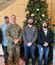 Seven volunteers (two in military camo) standing before a decorated Christmas tree with donation boxes and wrapped gifts.