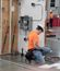 A man in an orange shirt and white hard hat kneels beside a competition electrical panel, working among tools and conduit pieces on the floor.