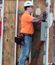 A man in an orange shirt and white hard hat stands at a wood-framed electrical mockup, using a cordless drill to install equipment.