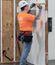 A man in an orange shirt and white hard hat measures across the top of a wall-mounted electrical panel with a tape measure.
