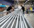 Two workers assemble metal conduit on a long fabrication table inside an industrial workshop.