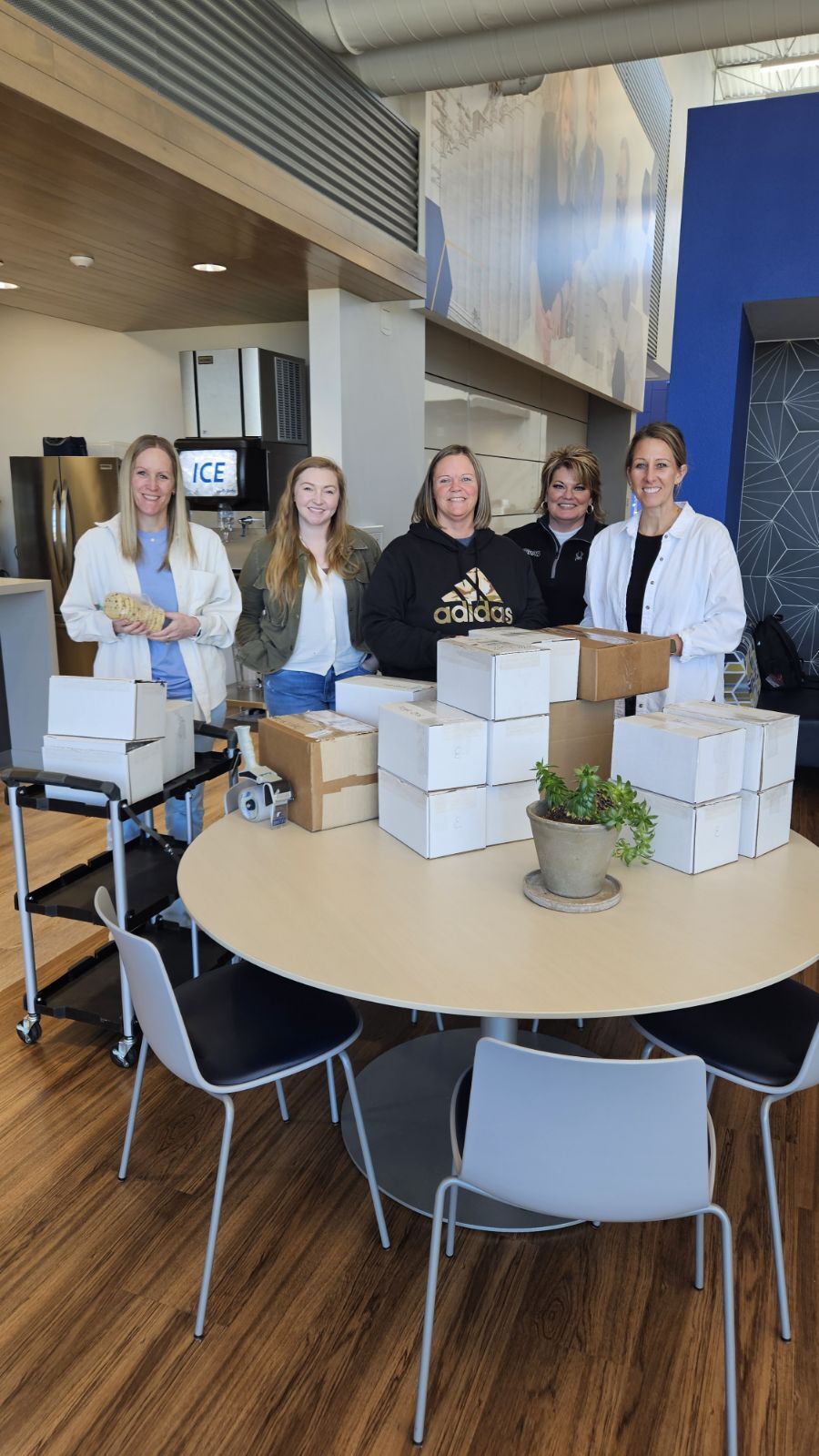 Five female employees standing behind table stacked with white boxes in modern break room, smiling and working together on packaging project