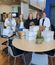 Five female employees standing behind table stacked with white boxes in modern break room, smiling and working together on packaging project