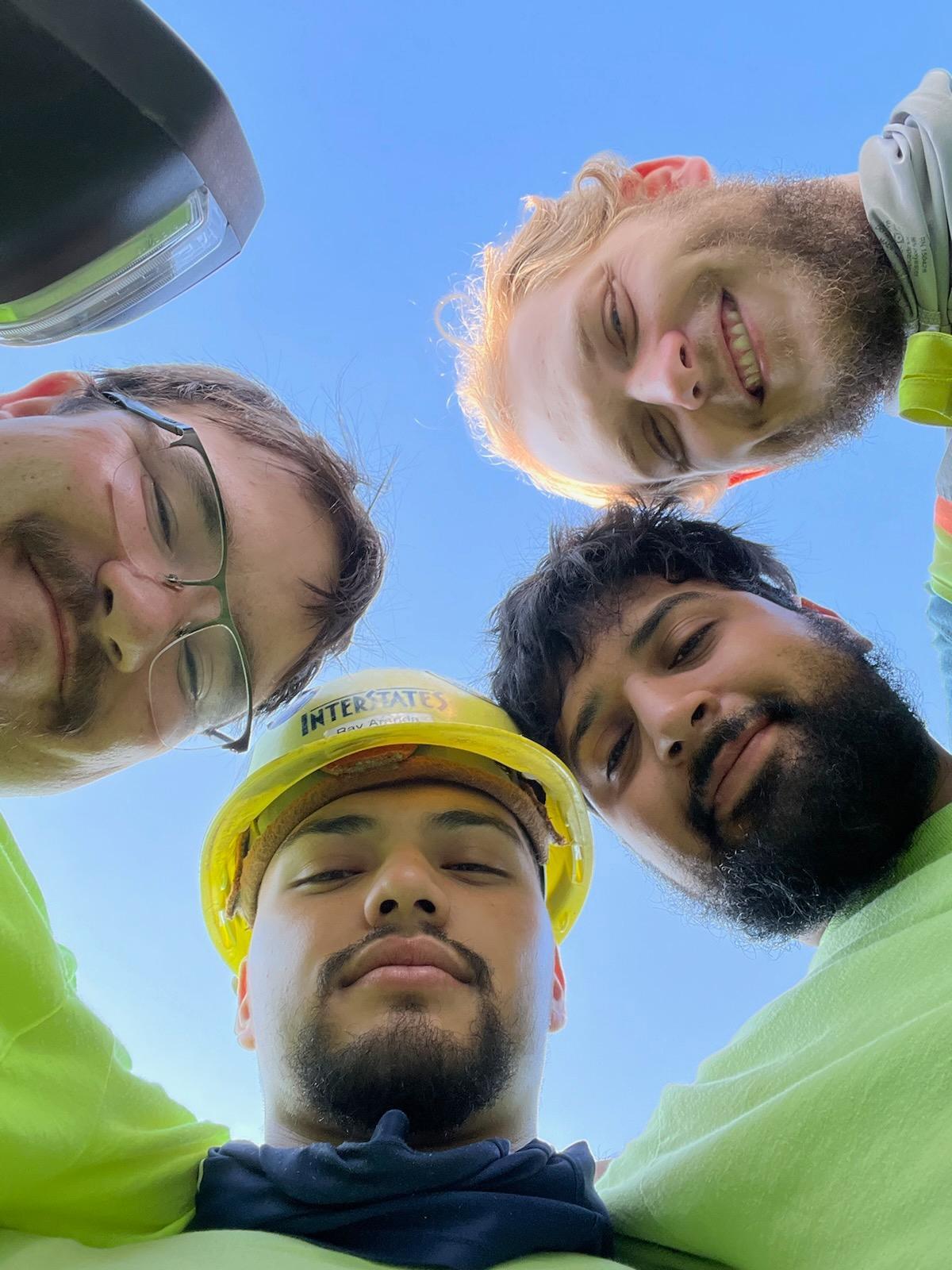 Low-angle selfie of four workers forming a circle looking down at camera, one wearing Interstate hard hat, with bright blue sky background