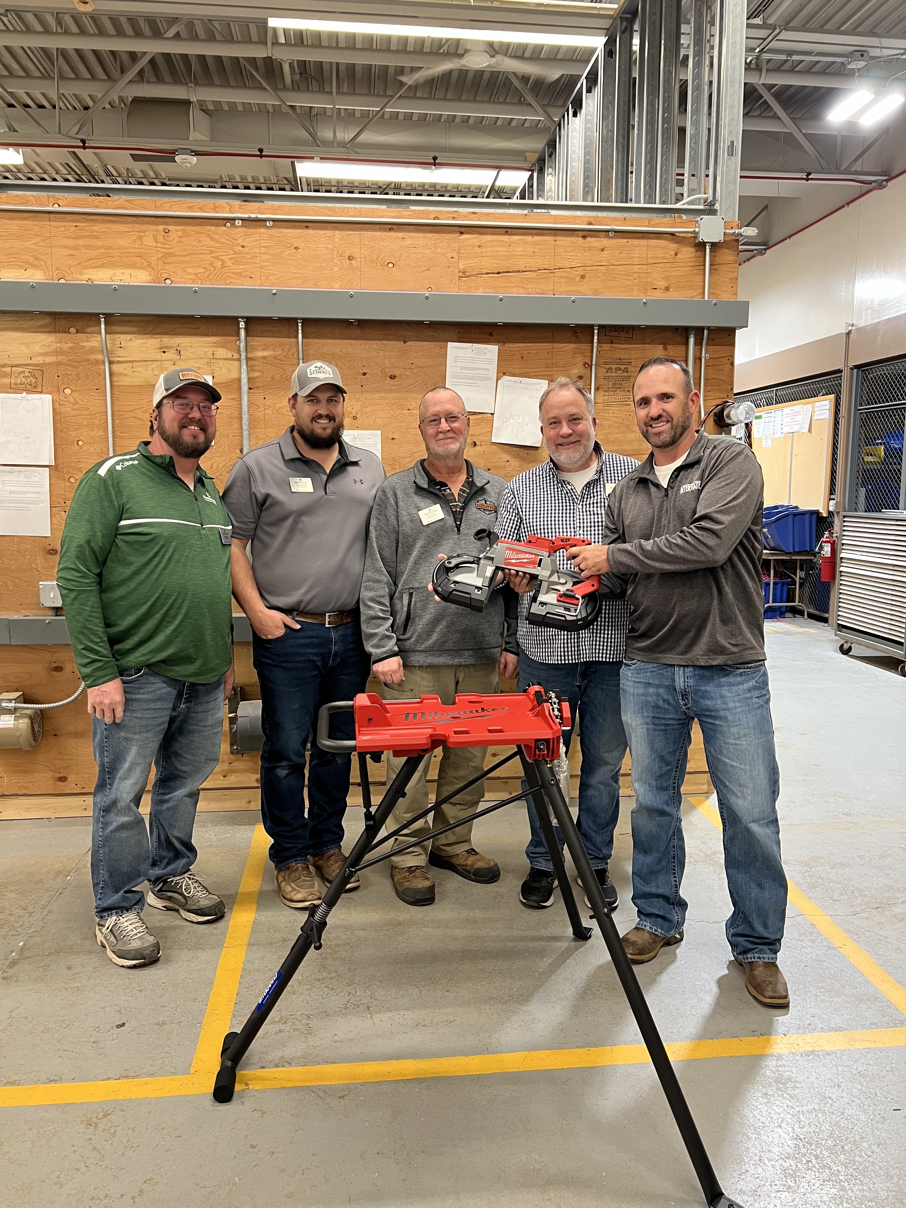 5 individuals standing in a shop holding a saw