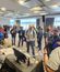 People stand and talk in a bright conference room during a team event, with a registration or display table in the foreground.