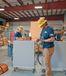 Two workers in hard hats and safety gear operate equipment in an industrial workshop with shelves and tools in the background.